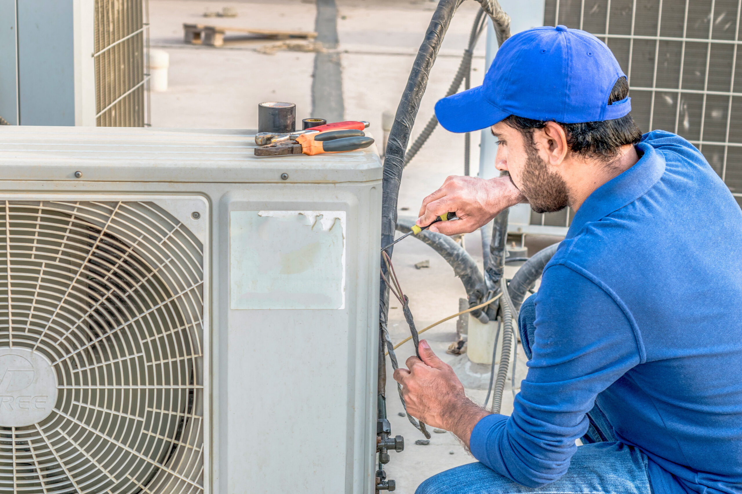 a professional electrician man is fixing a heavy duty unit of central air conditioning system by his tools on the roof top and wearing blue color of uniform and white cap HVAC Service Solutions Logo